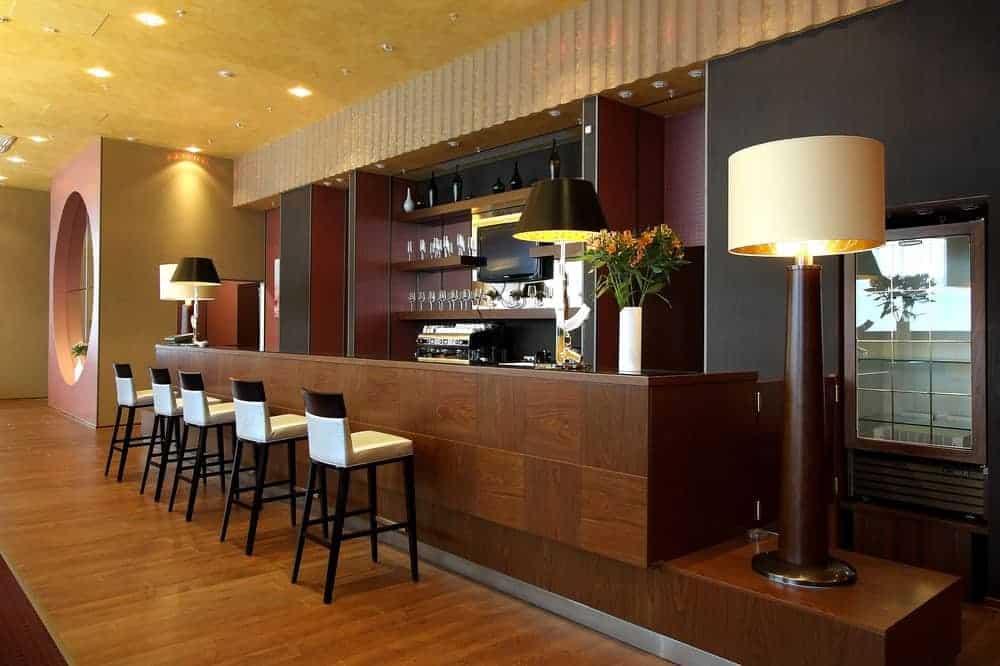  bar hotelowy z drewnianą blatem barowym, drewnianą podłogą, eleganckimi krzesłami barowymi i dużą lampą podłogową.